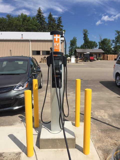 An electric vehicle (EV) charging station labeled 'EV Charging Only,' located in a parking lot between two cars. The station is surrounded by yellow bollards, with buildings, trees, and a clear blue sky in the background.